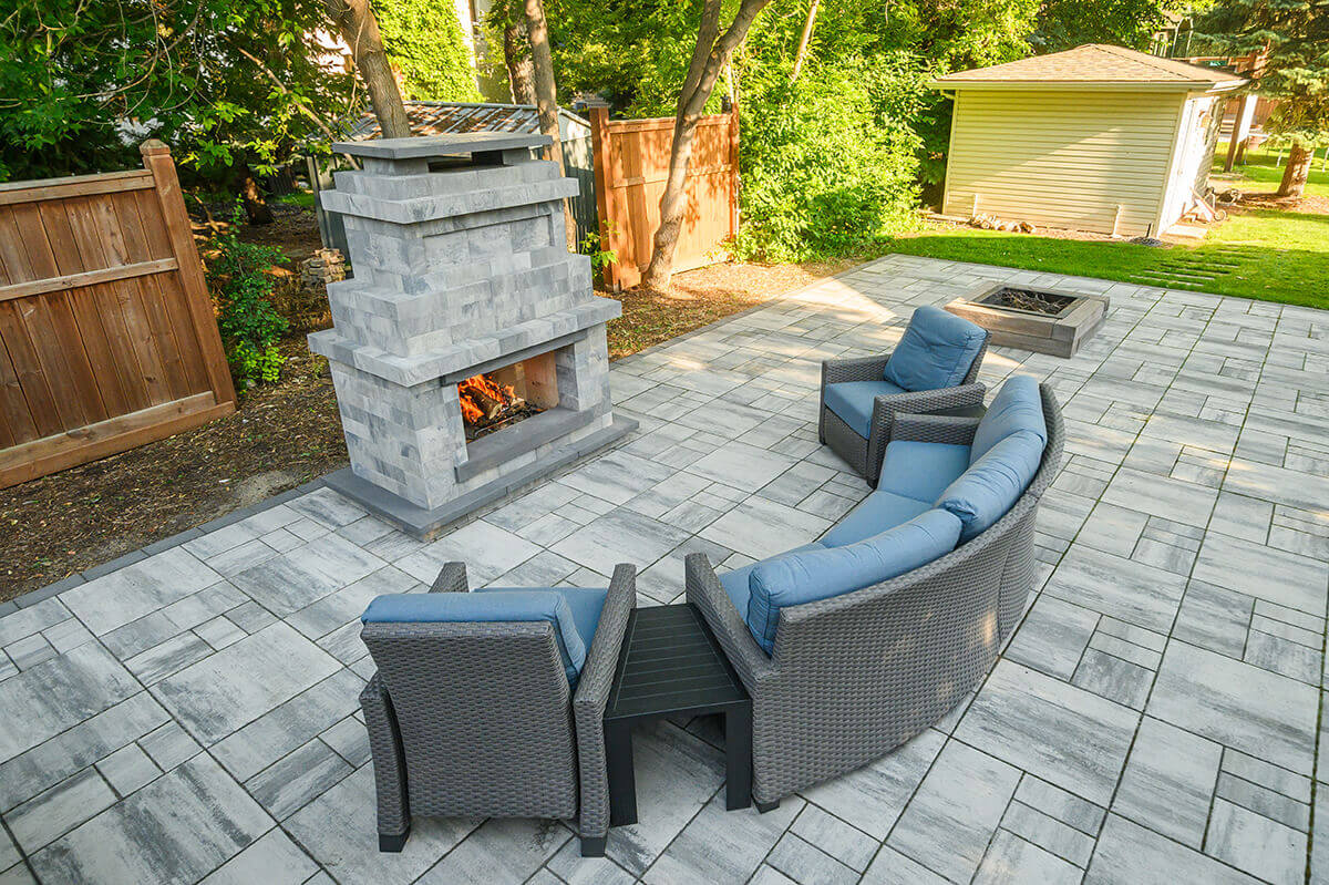 Expansive patio view showing the Caliber Stone Fireplace as the centrepiece of a contemporary seating arrangement with grey wicker furniture and curved blue cushions. Flames dance softly within the central hearth, built from multi-toned grey concrete blocks topped with charcoal stone accents. The surrounding patio features matching pavers in a seamless pattern that enhances the space’s cohesive look. The design encapsulates one kit built your way, offering a stylish, flexible solution for modern outdoor living.
