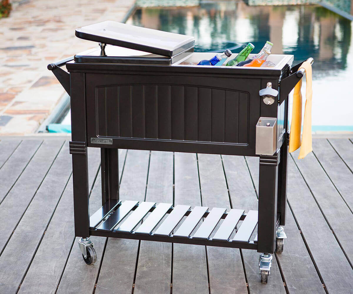 Outdoor setting featuring the Permasteel 80qt Furniture Style Cooler in black, positioned on a wooden deck beside a swimming pool. The cooler’s insulated basin is open and filled with colourful bottled drinks surrounded by ice. The design includes vertical panel-style embossing, a built-in bottle opener with a cap catcher, and a slatted lower shelf for storage. The glossy powder-coated steel surface reflects daylight, evoking a luxurious yet practical design for stylish summer entertaining.
