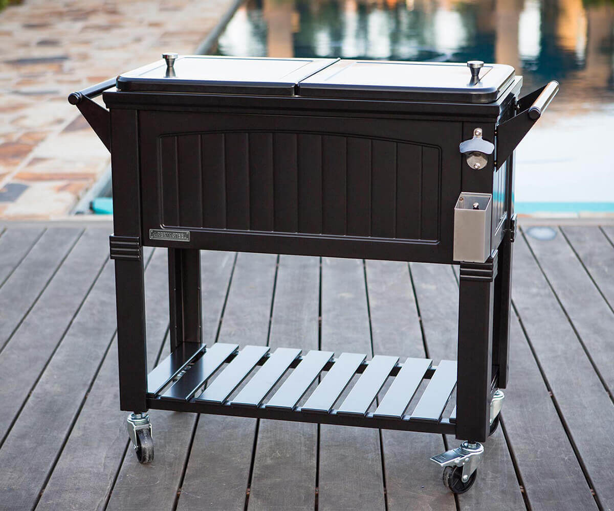 Front angled view of the Permasteel 80qt Furniture Style Cooler in black, placed on a wooden patio deck near a pool. The cooler’s dual-lid top is closed, and the surface reflects soft outdoor lighting. A bottle opener and matching cap catcher are attached to the right side, while a horizontal handle extends outward. The slatted shelf below offers added storage space, highlighting both decorative appeal and practicality.