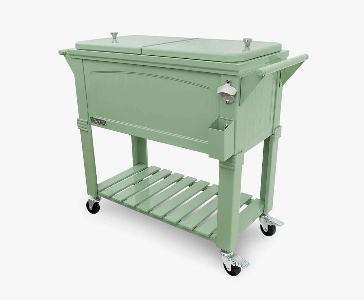 Angled product view of the Permasteel 80qt Furniture Style Cooler in soft sage green. The cooler has a rectangular shape with a raised decorative front panel, twin-lid top, and powder-coated steel frame. The structure includes large side handles, a built-in bottle opener with a cap catcher, and a slatted lower shelf supported by four lockable caster wheels. Its muted green colour and balanced proportions combine functionality with a refined vintage-inspired aesthetic.