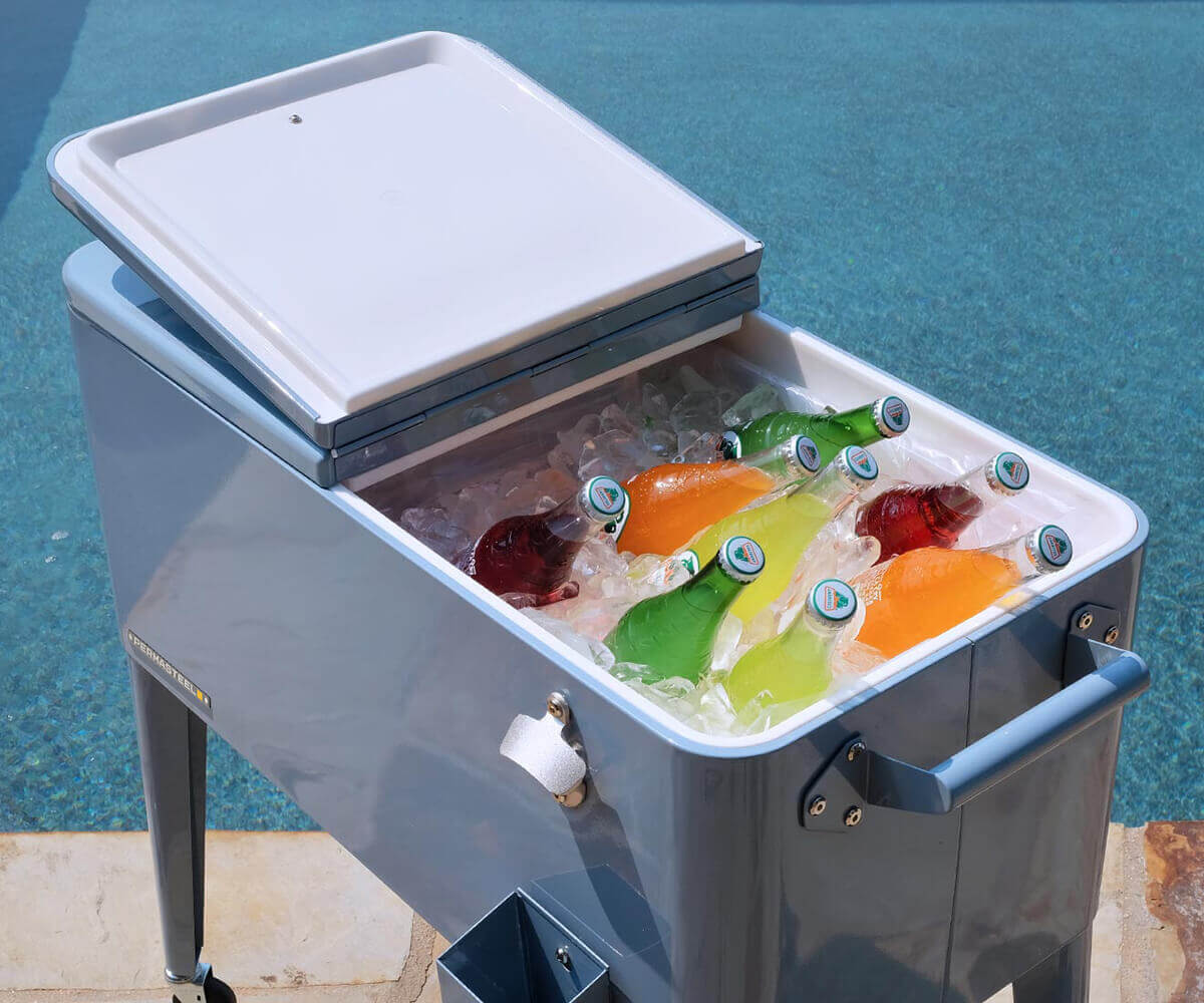 Close-up angled view of the Permasteel 80-quart rolling cooler in a glossy sky-blue finish. The cooler is filled with colourful glass bottles resting on crushed ice beside a bright swimming pool. The hinged split lid is partially open, revealing the insulated white interior. The steel handles, silver bottle opener, and matching cap catcher accentuate the cooler’s retro yet durable design, evoking a fun, refreshing summer atmosphere for outdoor entertaining.