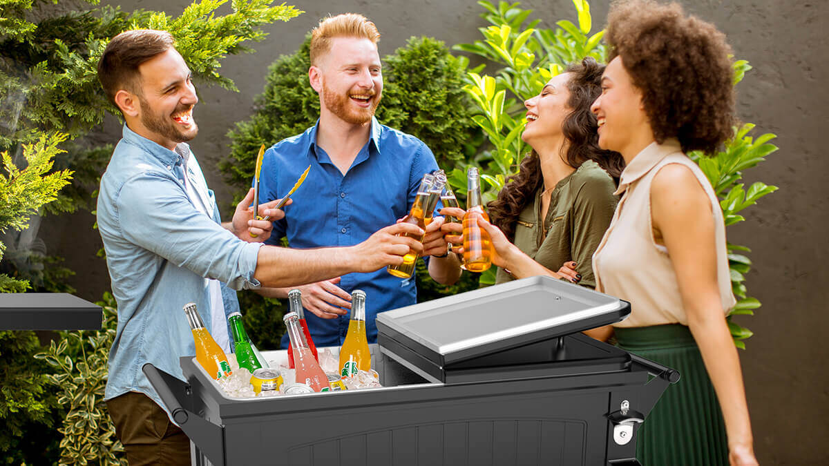 A cheerful group of four adults stands outdoors by a sleek black Permasteel cooler filled with ice and assorted colourful bottled drinks. The cooler features a split hinged lid, chrome bottle opener, and matching metal cap catcher. Two men and two women smile and laugh as they clink their drinks together, framed by lush green plants and soft natural sunlight. The cooler’s rectangular insulated basin is open on one side, showing bottles in vivid shades of orange, red, green, and yellow nestled in clear ice cubes. The outdoor setting and bright atmosphere evoke a lively summer gathering, capturing the cooler as a stylish and functional centrepiece for entertaining.