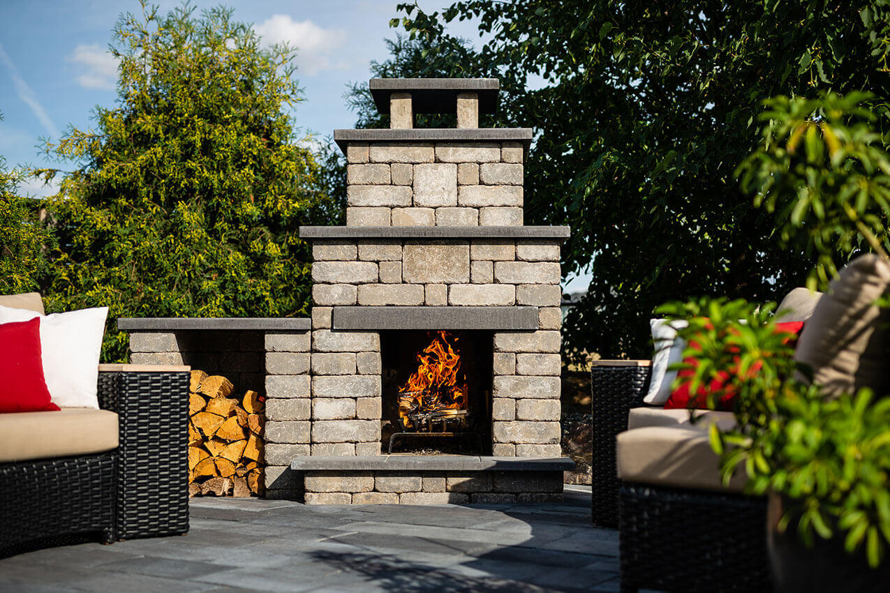 Cosy outdoor patio featuring the Quarry Stone Compact Fireplace framed by modern wicker seating with beige cushions and red accent pillows. The fireplace, made of light grey textured blocks with dark slate trim, stands against lush green foliage while a lively fire glows at its centre. Neatly stacked golden firewood fills the side compartment, enhancing its rustic elegance. This beautifully balanced design demonstrates how you can add a fireplace to any backyard, no matter the size, creating a welcoming retreat that feels expansive yet intimate.