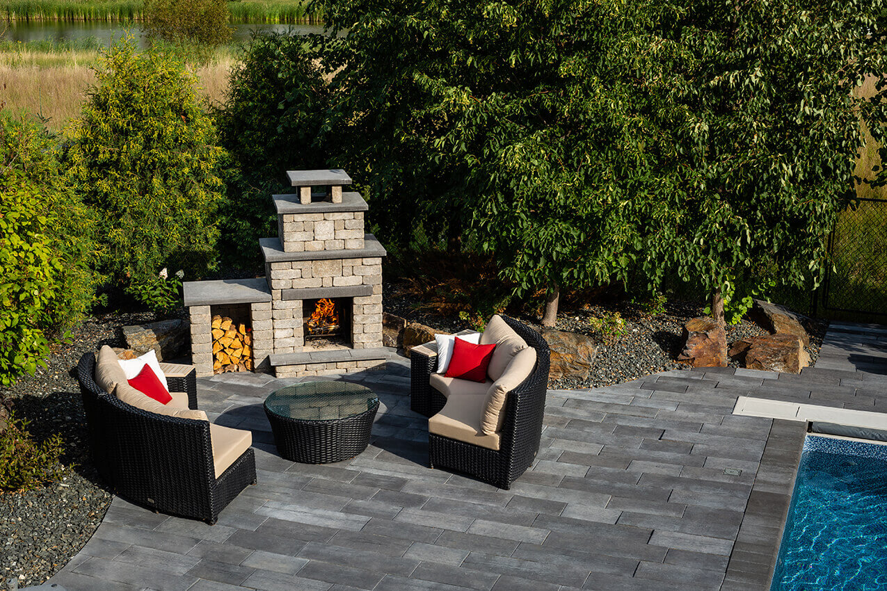 Aerial view of a serene backyard patio with the Quarry Stone Compact Fireplace positioned beside a bright blue swimming pool. Two curved wicker lounge chairs with red and white cushions frame the scene, while the fireplace’s light grey concrete structure with dark slate details emits a gentle orange flame. A small side compartment neatly stores logs, adding natural warmth to the modern layout. Perfect for patios where space is limited, this design proves you can add a fireplace to any backyard, no matter the size, without compromising elegance or comfort.
