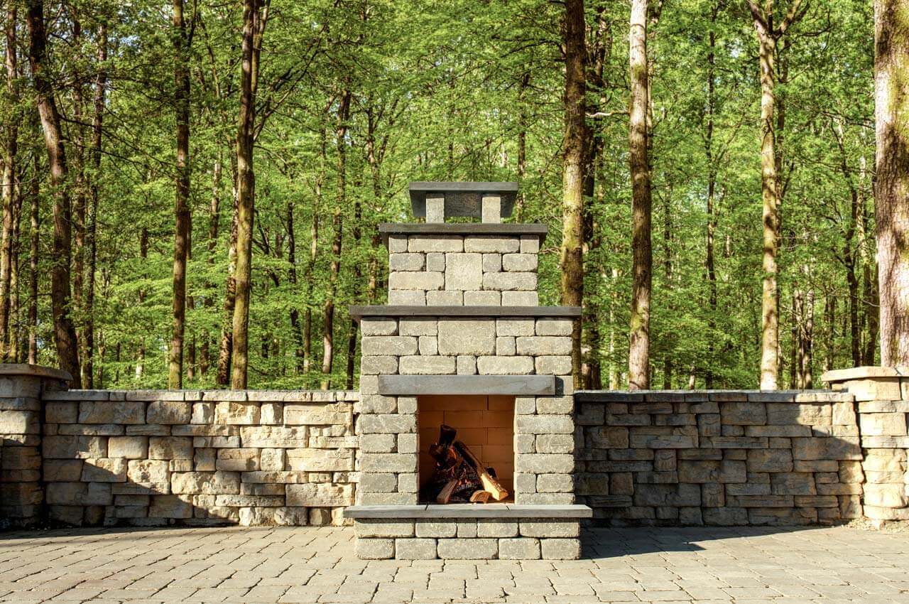 Rustic forest patio showcasing the Quarry Stone Compact Fireplace built against a tall stone retaining wall. The structure, made from light grey and beige masonry blocks with dark slate shelves and chimney top, houses a neatly arranged pile of logs ready for lighting. Surrounded by dense green woodland and dappled sunlight, the fireplace appears perfectly integrated into nature. It demonstrates how thoughtful design allows you to add a fireplace to any backyard, no matter the size, blending timeless stone craftsmanship with a peaceful outdoor atmosphere.