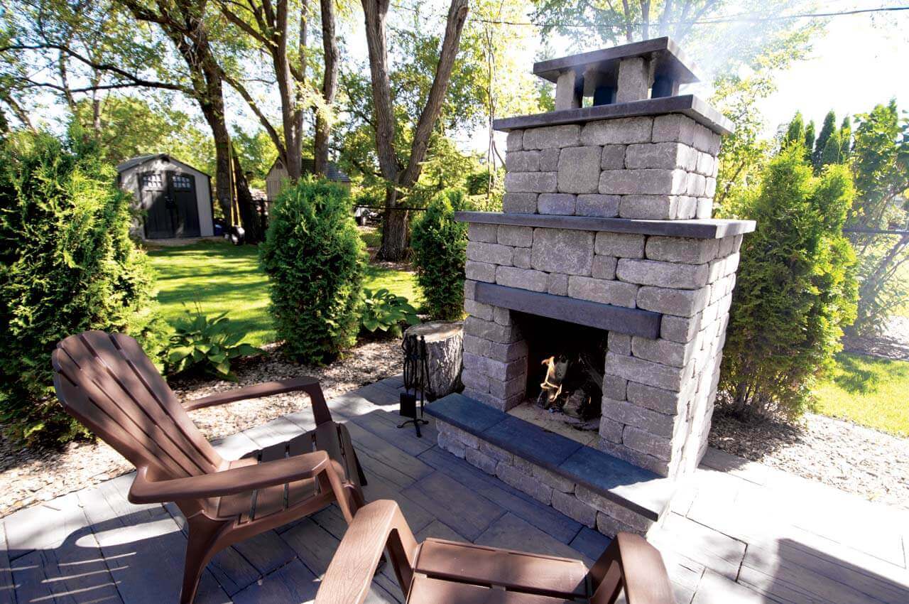 Inviting backyard setting with the Quarry Stone Compact Fireplace placed near brown Adirondack chairs on a neatly paved patio. Built from textured grey concrete blocks with dark slate trim, the fireplace radiates gentle warmth as a small flame flickers within the hearth. Surrounded by manicured shrubs and filtered sunlight through tall trees, the space feels open yet intimate. This clever, compact design makes it easy to add a fireplace to any backyard, no matter the size, turning even a modest garden into a comfortable outdoor retreat.
