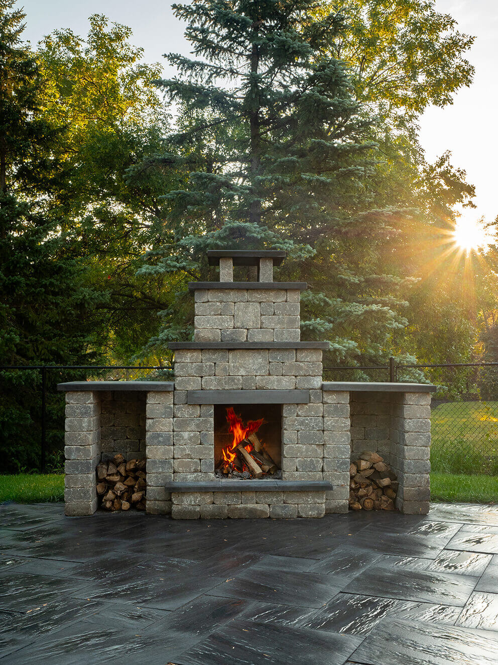 Evening view of the Quarry Stone Compact Fireplace standing on a polished dark grey herringbone patio, framed by soft sunlight filtering through pine trees. The fireplace features light grey stone blocks contrasted with charcoal slate mantels and a capped chimney, with neatly stacked logs in side compartments. A bright orange fire flickers in the centre, casting warm light across the sleek stonework. Ideal for smaller yards and patios, it embodies the concept of adding a fireplace to any backyard, no matter the size, for year-round outdoor enjoyment.