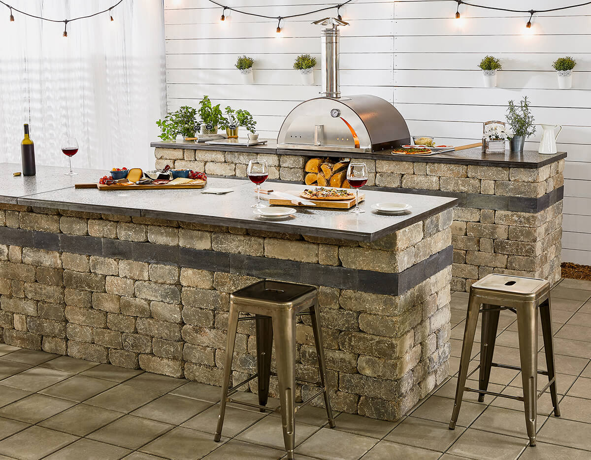 Inviting modern outdoor kitchen scene featuring the Quarry Stone Compact Pizza Oven integrated into a rustic stone island beneath warm café-style string lights. The oven’s hammered copper dome and stainless steel chimney contrast with the rough-textured grey and beige stone base. The sleek countertop displays freshly baked pizzas, wine glasses, and charcuterie boards, capturing a lively yet relaxed atmosphere. The entire setup showcases how this compact pizza oven merges style and functionality, offering a refined space for cooking gourmet meals in any backyard setting.