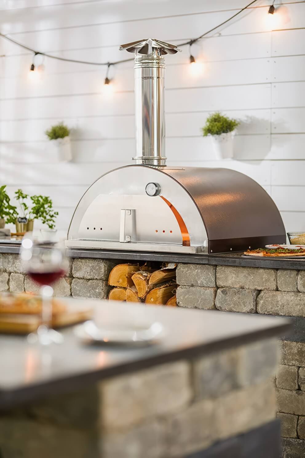 Close side view of the Quarry Stone Compact Pizza Oven highlighting the hammered copper dome and stainless steel chimney gleaming beneath soft ambient lighting. The oven rests on a robust stone countertop built from weathered beige blocks accented by a dark charcoal horizontal band. Below, neatly stacked split firewood fills the lower storage cavity. A glass of red wine and blurred culinary items in the foreground subtly frame the scene, reflecting the oven’s role as both a culinary centrepiece and an elegant focal point for outdoor gatherings.