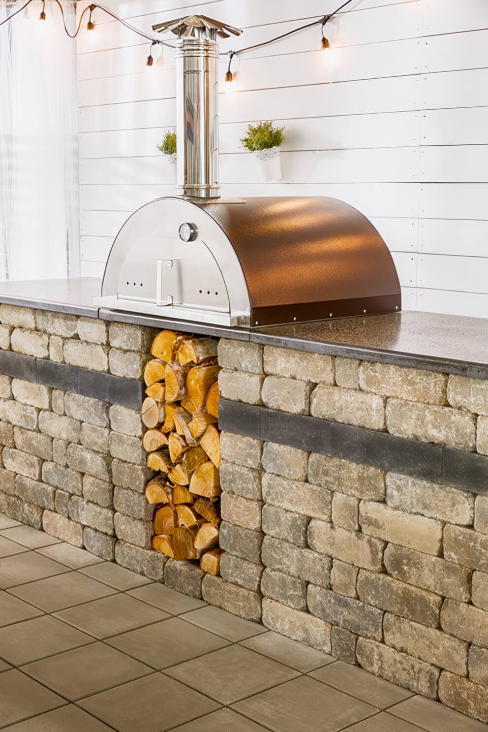 Front view of the Quarry Stone Compact Pizza Oven unit showing its distinctive arched hammered copper dome paired with a polished stainless steel oven front and chimney. The oven sits atop a rectangular stone counter constructed from light grey and taupe-coloured blocks with dark charcoal accents. A vertical storage space below the oven holds a column of stacked split firewood, providing both convenience and symmetry. Decorative string lights and wall-mounted plants complete the sophisticated yet functional design, celebrating craftsmanship and versatility in outdoor kitchen setups.