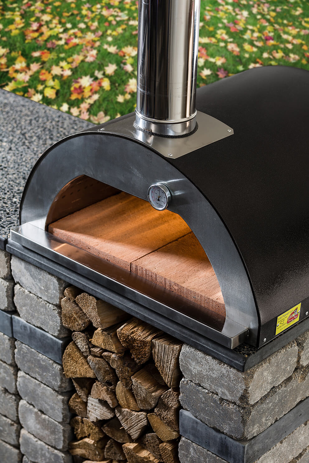 Detailed close-up of the Quarry Stone Compact Pizza Oven showing the black dome version of the Nonno Peppe oven with a brushed stainless steel front and built-in thermometer. The open oven cavity reveals terracotta-coloured firebricks that form the cooking surface. The oven is mounted on a sturdy stone base made from natural grey bricks with dark slate bands, and firewood is neatly stored below. The surrounding area features speckled countertop material and autumn leaves scattered on green grass, capturing the oven’s charm and efficiency for year-round outdoor cooking.