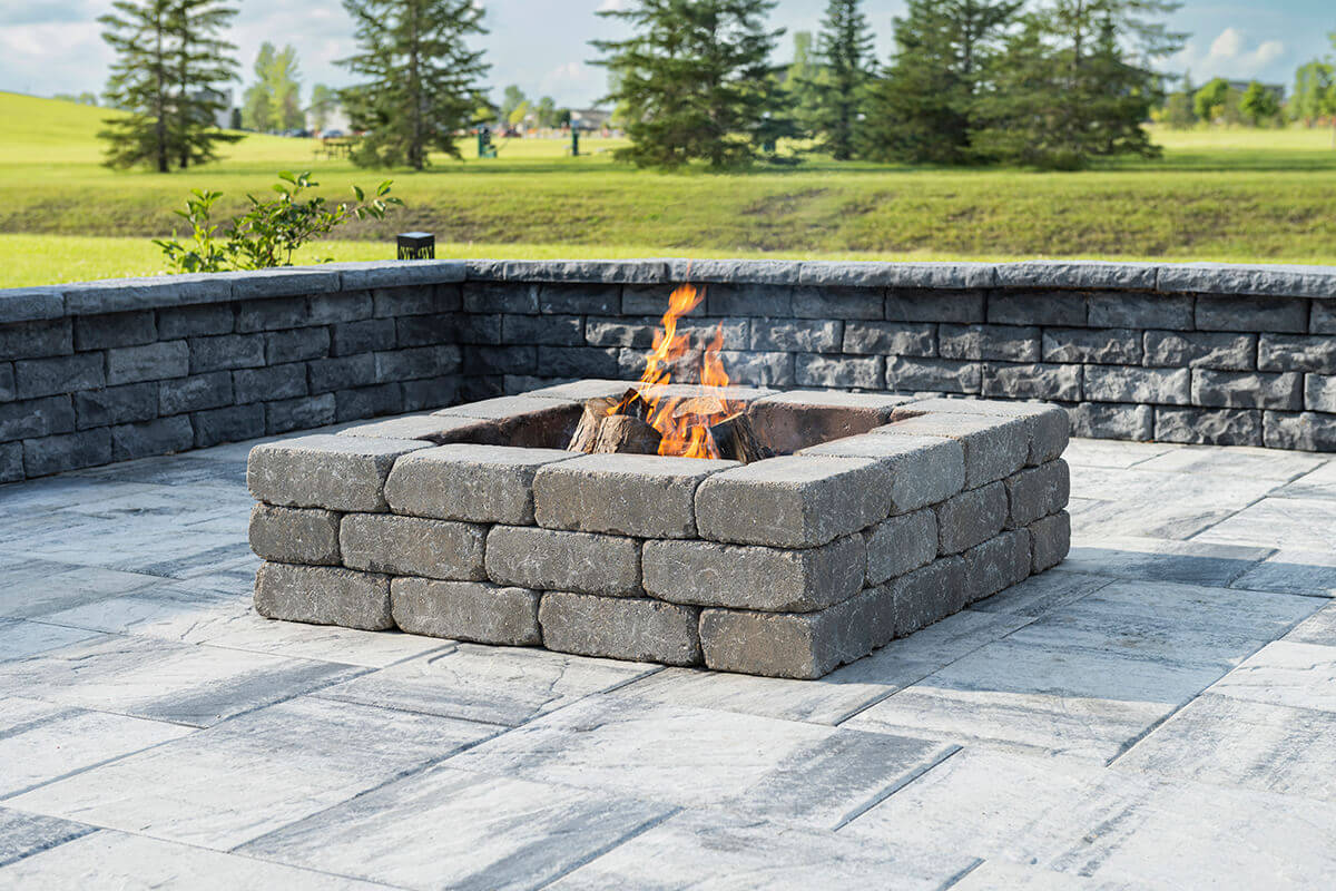 A square Quarry Stone Fire Pit positioned on a large, light-grey paver patio bordered by matching retaining walls. The fire pit is built from stacked, tumbled concrete blocks in a deep slate-grey tone with weathered texture and faint white accents. Inside, orange flames rise above blackened firewood, giving off a gentle glow. Behind the setup, lush green grass and tall evergreen trees complete the serene outdoor backdrop. The blend of rugged materials and natural tones enhances the scene—an inviting space designed to complement your space with a rustic fire pit.