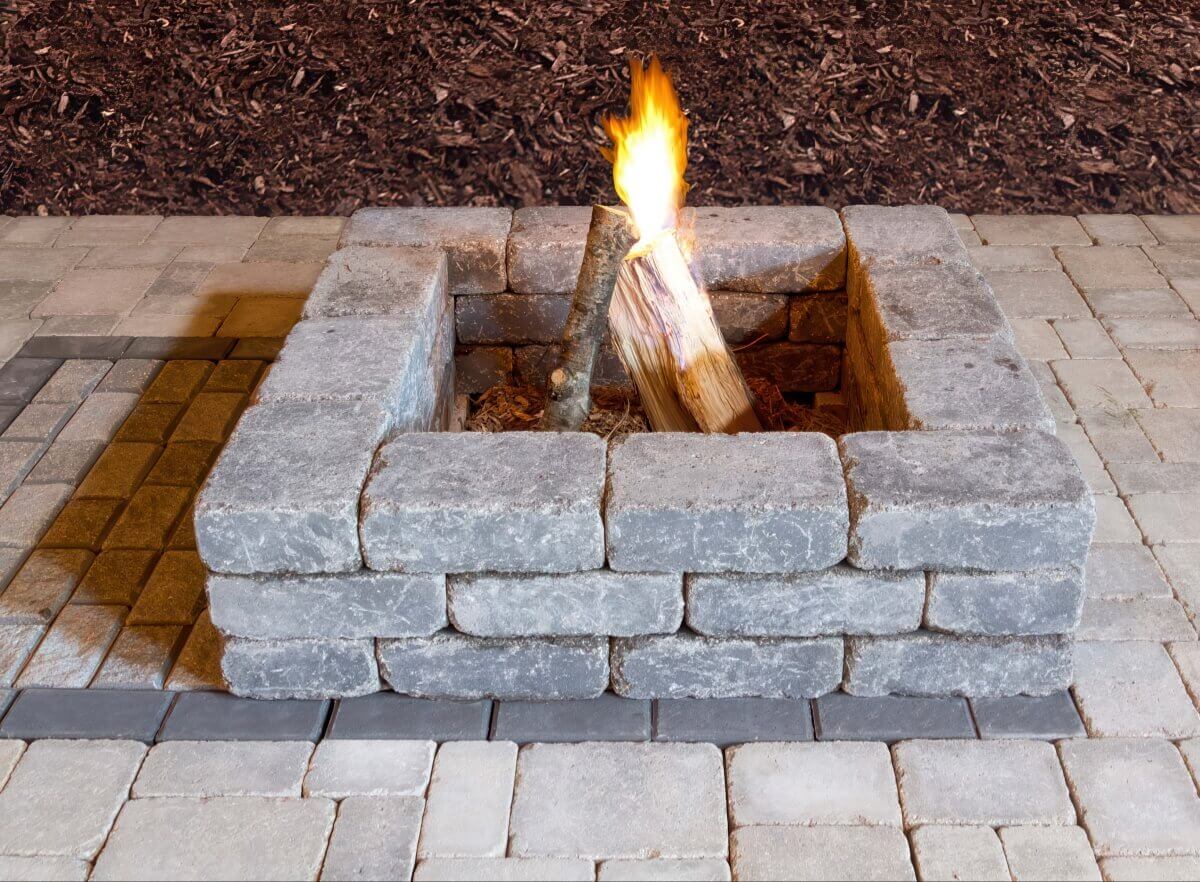 Close-up view of a Quarry Stone Fire Pit resting on interlocking pavers in varied tan and charcoal shades. The square structure consists of thick, rounded concrete blocks arranged in three sturdy tiers, featuring rough stone-like faces with light surface marbling. A single upright log burns brightly at the centre, casting warm yellow and orange hues across the textured stone edges. The pit’s compact form and handcrafted appearance express rustic charm, seamlessly complementing your space with a rustic fire pit that brings comfort and visual appeal.