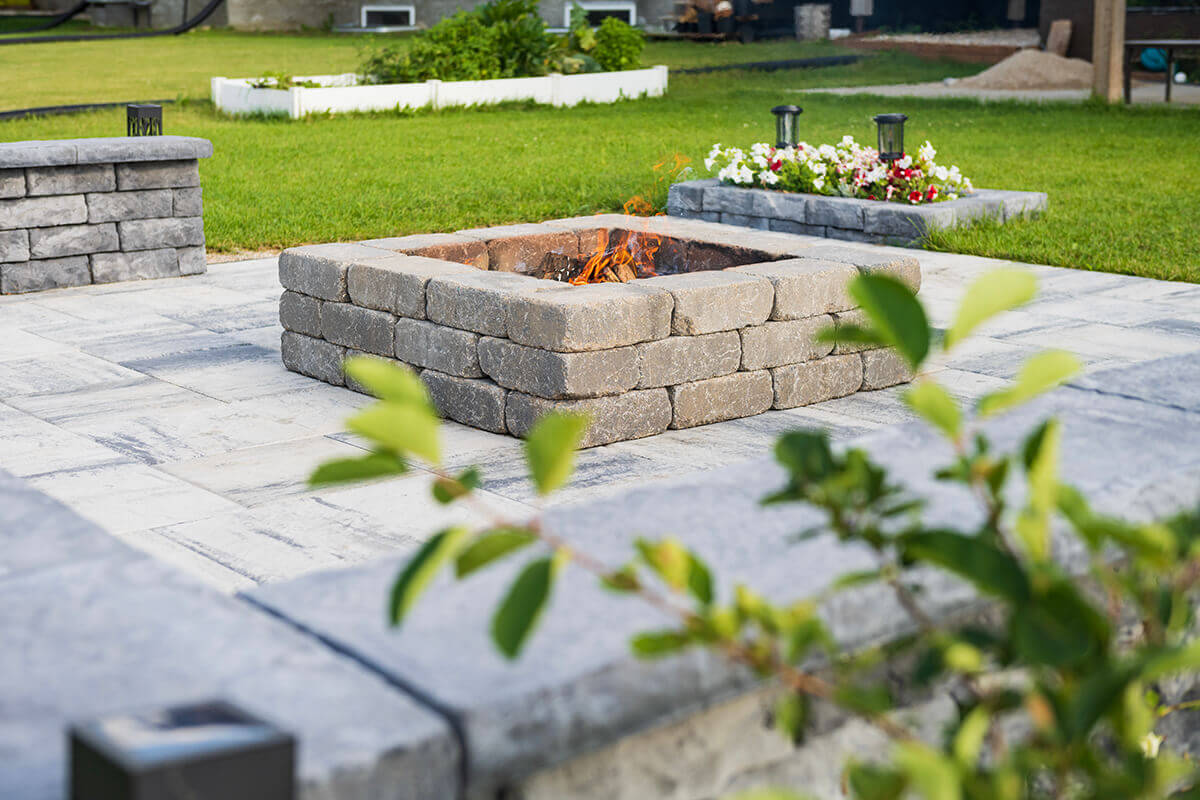 Backyard patio scene showing a Quarry Stone Fire Pit made from uniform, tumbled concrete blocks in a weathered charcoal-grey tone with soft white flecking. The pit’s square layout is neatly aligned on pale patterned patio stones surrounded by green grass and a small flower bed blooming with white and red blossoms. The firewood within burns gently, the orange flames flickering against the subtle texture of the stone. This elegant yet rugged design merges durability with natural charm—crafted to complement your space with a rustic fire pit for all-season enjoyment.