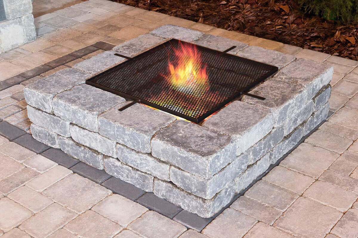 Evening patio view of a Quarry Stone Fire Pit featuring a black metal mesh spark screen over the open flame. The square pit is built from rough-textured, tumbled concrete blocks in a cool grey tone with lighter streaks accentuating its stone-like surface. Beneath the soft orange and red firelight, the material’s contours appear slightly glossy. The pit rests on a paved surface of beige and grey bricks, surrounded by mulch and garden edging. The mix of function and rustic style effortlessly complements your space with a rustic fire pit that radiates warmth and safety.