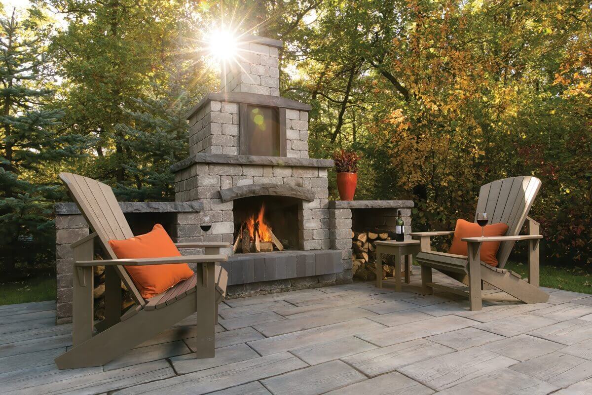Tranquil outdoor patio scene featuring the Quarry Stone Fireplace surrounded by tall green trees and soft evening sunlight. The fireplace, constructed from light grey concrete blocks with darker slate trim, burns with bright orange flames beneath an arched lintel. Two wide armchairs with orange cushions and glasses of red wine create a warm and inviting atmosphere. The realistic stone texture and balanced design capture the spirit of living designed to entertain outdoors, turning this patio into a sophisticated woodland retreat.