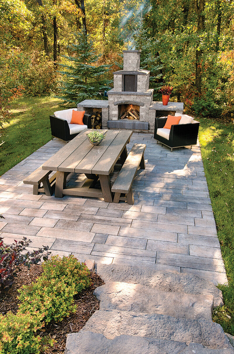 Wide overhead view of the Quarry Stone Fireplace as the centrepiece of a forest-edge patio furnished with a rustic wooden picnic table, matching benches, and cushioned lounge chairs. The fireplace’s sturdy grey stone form with dark slate details radiates warmth from a small fire glowing inside its arched firebox. Smoke gently rises into the crisp autumn air surrounded by golden foliage. The arrangement illustrates how this fireplace is designed to entertain outdoors, bringing people together in a natural, relaxing space.