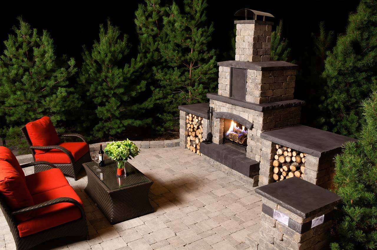 Night-time patio illuminated by the soft orange glow of the Quarry Stone Fireplace. Built from stacked grey blocks with charcoal-coloured caps and mantle ledges, it stands elegantly against a backdrop of dark evergreen trees. The fire crackles brightly within the arched firebox, casting reflections across the light stone patio. Two rattan lounge chairs with bold red cushions and a glass-top table sit nearby, evoking comfort and warmth. Every detail of this space showcases a design meant to entertain outdoors with style and ease.