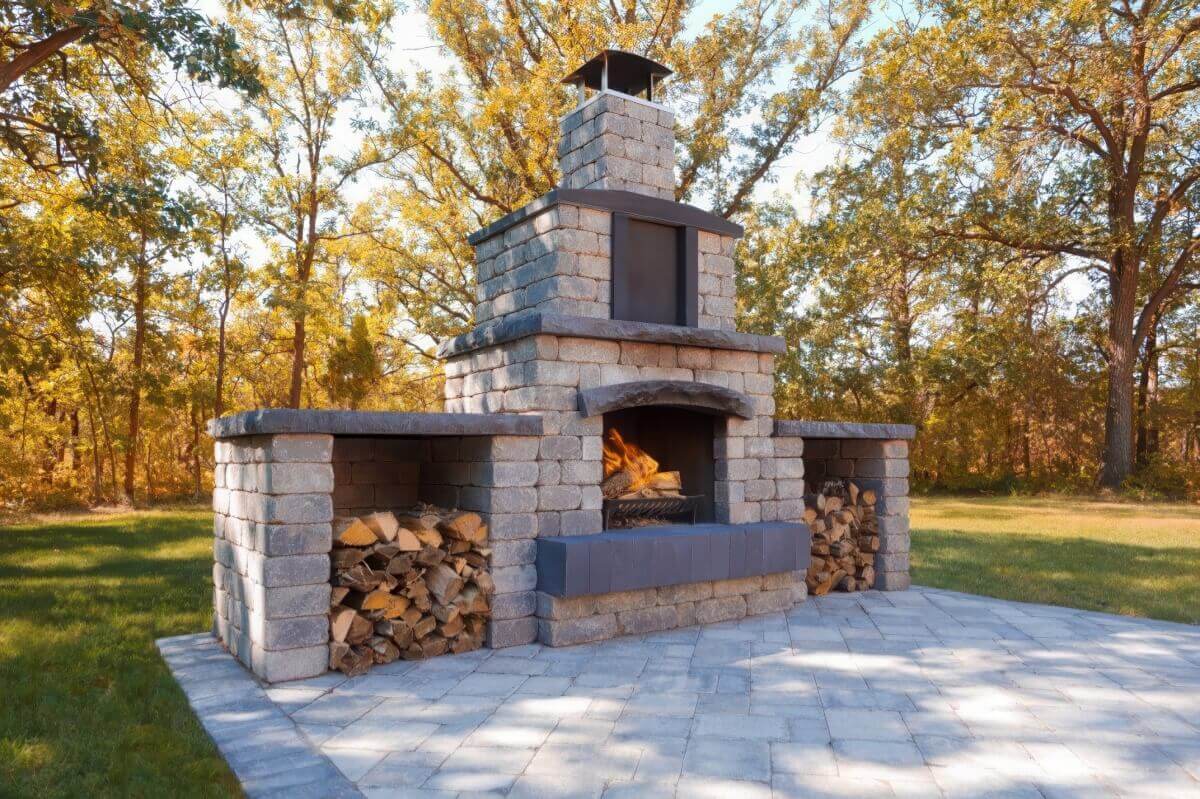 Daytime view of the Quarry Stone Fireplace set on a pale stone patio surrounded by sun-lit trees. The fireplace features multi-toned grey masonry blocks, a gently curved arched opening, and wide storage shelves filled with split firewood on each side. A black metal chimney cap crowns the structure, complementing the slate-coloured trim. The design embodies a perfect blend of strength and elegance, created to entertain outdoors while enhancing the natural beauty of open-air living spaces.