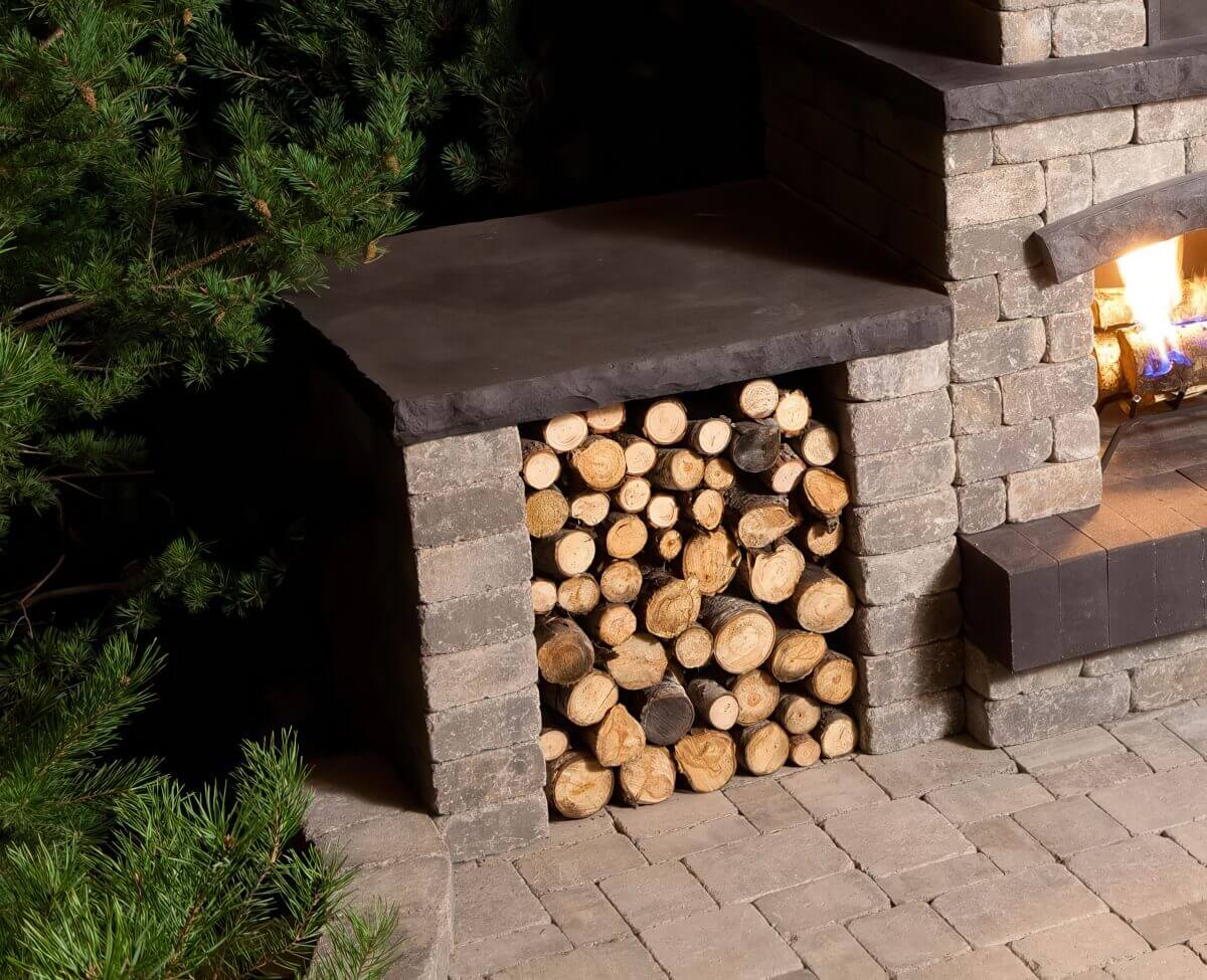 Close-up of the left-side log storage compartment of the Quarry Stone Fireplace, revealing stacked firewood arranged beneath a smooth charcoal-coloured stone ledge. The texture of the light grey stone blocks contrasts with the natural wood grain and the soft glow of firelight nearby. The scene captures the rugged craftsmanship and thoughtful functionality of a fireplace designed to entertain outdoors, adding both warmth and rustic sophistication to backyard gatherings.