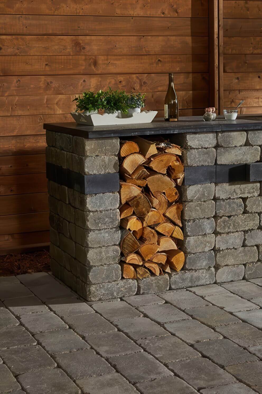 Front view of the Quarry Stone Storage Unit incorporated into a rustic outdoor kitchen setup against a cedar-panelled wall. The unit is constructed from rough-faced grey and tan stone blocks featuring a dark horizontal accent band and topped with a sleek grey countertop. Split logs are neatly arranged in the open centre cavity beneath potted herbs and a bottle of wine on the counter. The image highlights the versatility of the storage unit as a functional and decorative element that complements outdoor living areas.