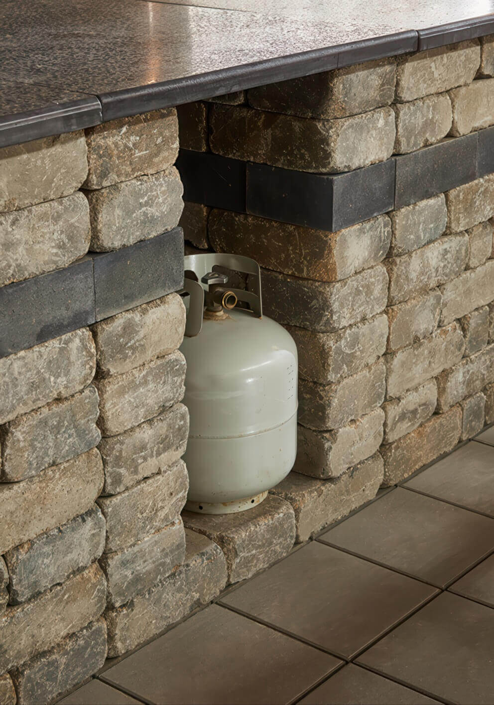 Close-up of the Quarry Stone Storage Unit beneath a speckled stone countertop, showcasing a propane tank neatly positioned in the recessed centre compartment. The stacked concrete blocks display a natural mix of grey and beige tones with a single dark charcoal band running horizontally for contrast. The scene emphasises the unit’s multipurpose design, ideal for concealing propane tanks or storing outdoor kitchen items while maintaining a cohesive and refined stone aesthetic.