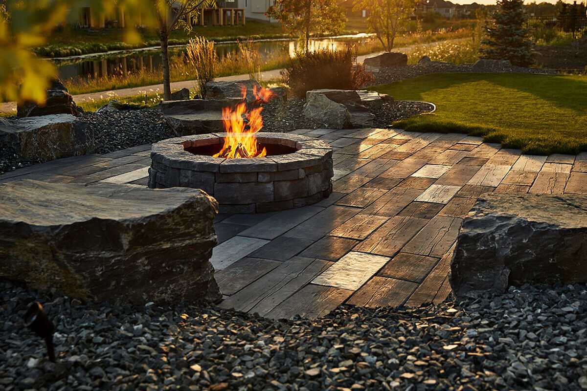 An inviting backyard patio at sunset featuring the Rosetta® Belvedere Fire Pit surrounded by rugged natural boulders and a curved lawn edge. The round fire pit is built from rough-textured precast concrete blocks that mimic hand-cut natural stone, with varying tones of deep grey and charcoal. Bright orange flames rise from stacked firewood in the centre, casting reflections across the long, rectangular paving stones that resemble weathered wood planks. The scene’s warm golden light and organic textures embody the feeling of unwinding outdoors — the perfect example of how you can settle in around the Rosetta Belvedere Fire Pit, generously sized and ready for a summer of backyard serenity.