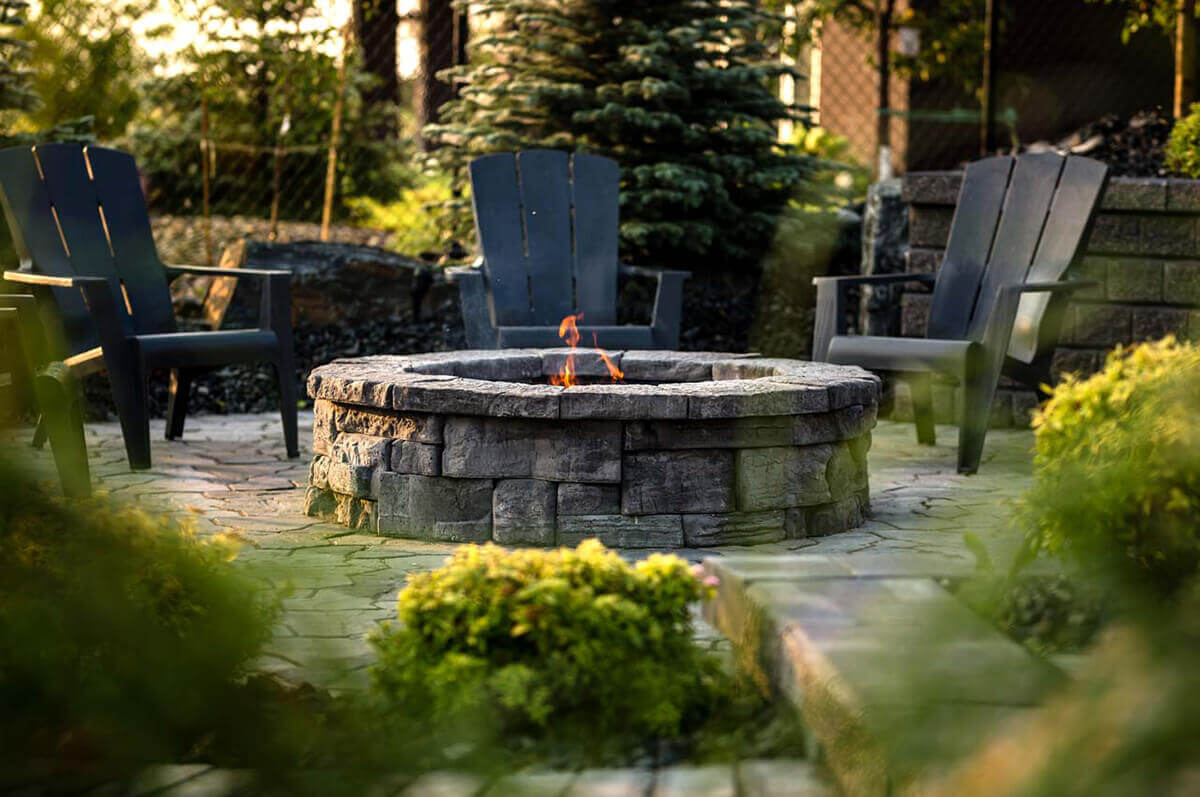 A peaceful garden setting framed by lush greenery with the Rosetta® Belvedere Fire Pit at its centre. The circular structure is made of stacked grey-stone blocks with a rough, layered texture and subtle colour variations that resemble natural quarry rock. Inside, faint orange flames flicker within a black steel ring, reflecting softly on the surrounding pavers. Four dark-charcoal Adirondack chairs encircle the pit in a semicircle, positioned over a flagstone patio bordered by evergreens and shrubs. Gentle morning light filters through, showing how the Belvedere Fire Pit’s natural-stone finish and generous size create a comfortable gathering place for backyard serenity.