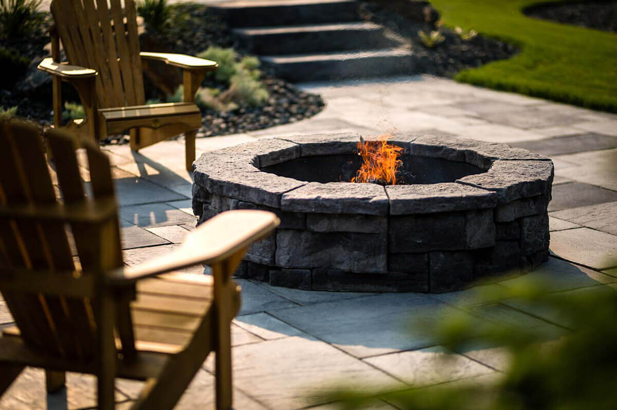 Sunlit outdoor patio featuring the Rosetta® Belvedere Fire Pit surrounded by two golden-cedar Adirondack chairs. The fire pit’s circular design is crafted from rugged grey-stone concrete blocks that mirror the look of hand-chiseled rock. A black steel liner holds a softly glowing orange flame at its centre. The large rectangular patio pavers around it are arranged neatly, leading the eye toward manicured green grass and garden steps beyond. The combination of sunlight, natural textures, and inviting warmth captures how the Belvedere Fire Pit invites you to settle in for a summer of backyard serenity.