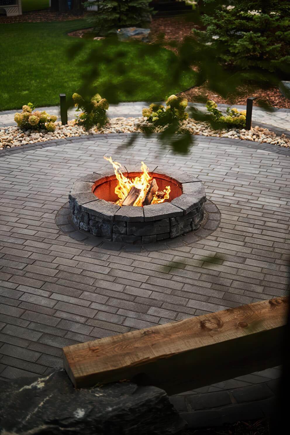Top-down view of a circular stone patio showcasing the Rosetta® Belvedere Fire Pit at its heart. The pit is built from rugged grey blocks, each detailed with deep grooves and weathered stone textures that create the look of natural rock. Inside the fire pit, logs burn brightly with golden flames that contrast against the dark, brick-patterned pavers. Around the circular patio are smooth river rocks, low hydrangea bushes, and manicured green grass. The soft evening light and careful arrangement of textures evoke a calm, inviting mood, illustrating how the Belvedere Fire Pit transforms any outdoor space into a serene summer retreat.