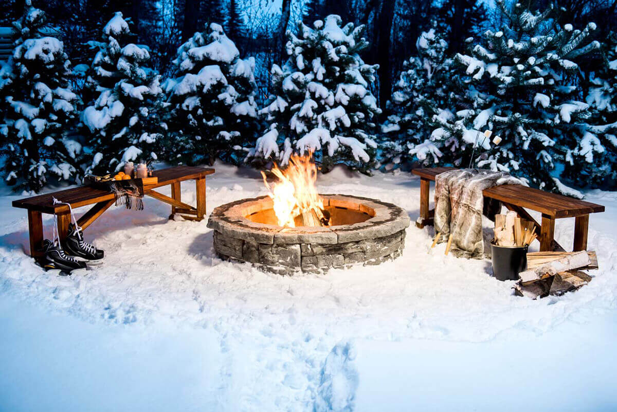 A picturesque winter evening scene showing the Rosetta® Belvedere Fire Pit glowing warmly amid freshly fallen snow. Surrounded by snow-covered evergreen trees, the fire pit’s round, hand-carved-look grey-stone blocks contrast against the bright white snow. Flames rise high from the centre, lighting up two rustic wooden benches positioned opposite each other. One bench has a faux-fur blanket, a metal pail of firewood, and roasting sticks leaning nearby; the other holds a pair of black ice skates and mugs of hot cocoa. The glowing fire against the cool winter backdrop beautifully expresses how the Belvedere Fire Pit invites you to settle in for backyard serenity all year round.
