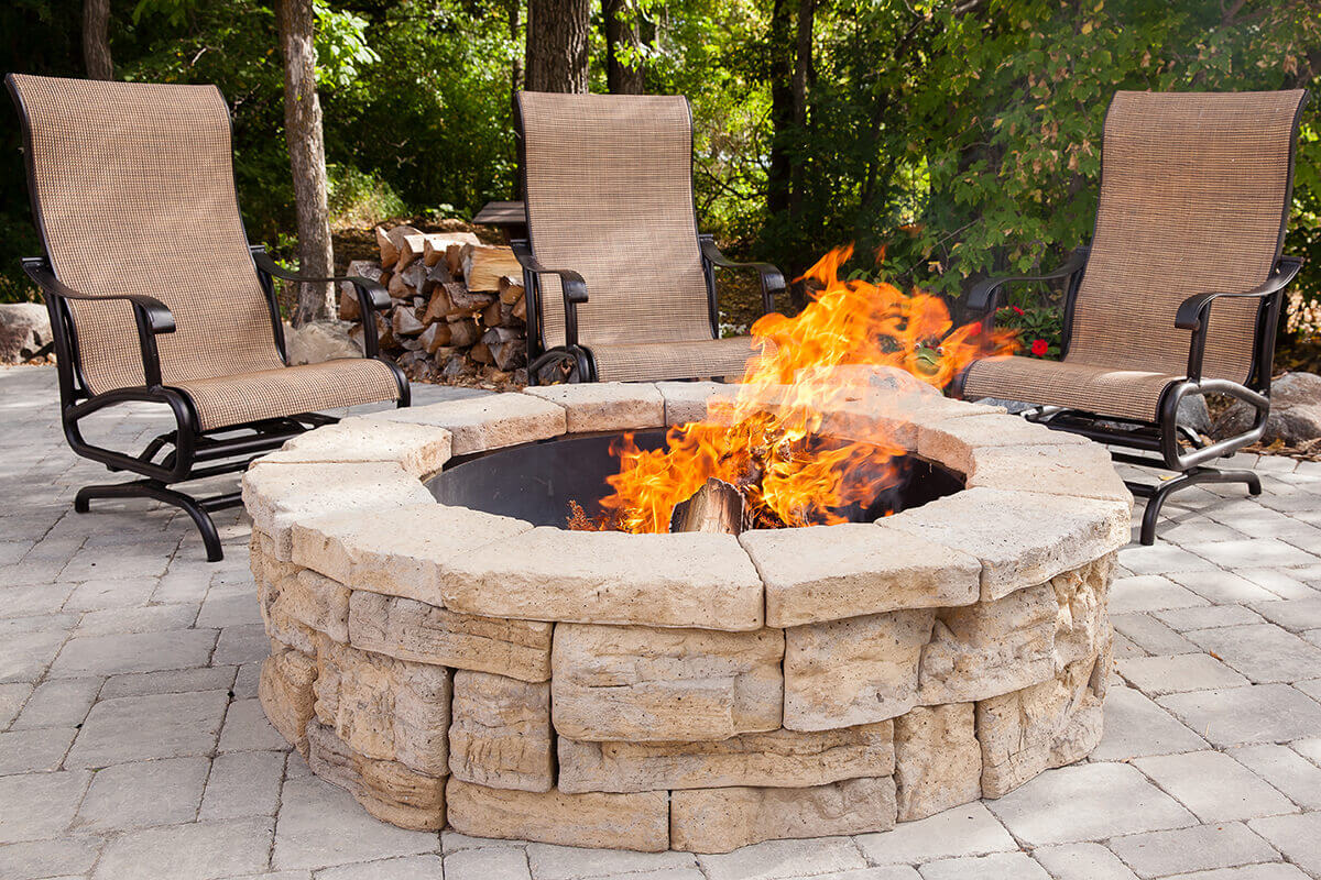Daytime patio scene surrounded by tall trees and natural stone landscaping featuring the Rosetta® Belvedere Fire Pit as its focal point. The circular pit is crafted from beige and tan stone-textured concrete blocks that replicate natural limestone with layered surfaces and subtle shading. A black steel insert lines the inner ring, holding burning logs and lively orange flames. Three high-backed patio chairs with woven tan fabric and curved metal frames surround the pit on a paved stone surface, with a neat stack of firewood resting nearby. The natural tones and generous proportions of the Belvedere Fire Pit perfectly capture the feeling of summer relaxation and backyard serenity.