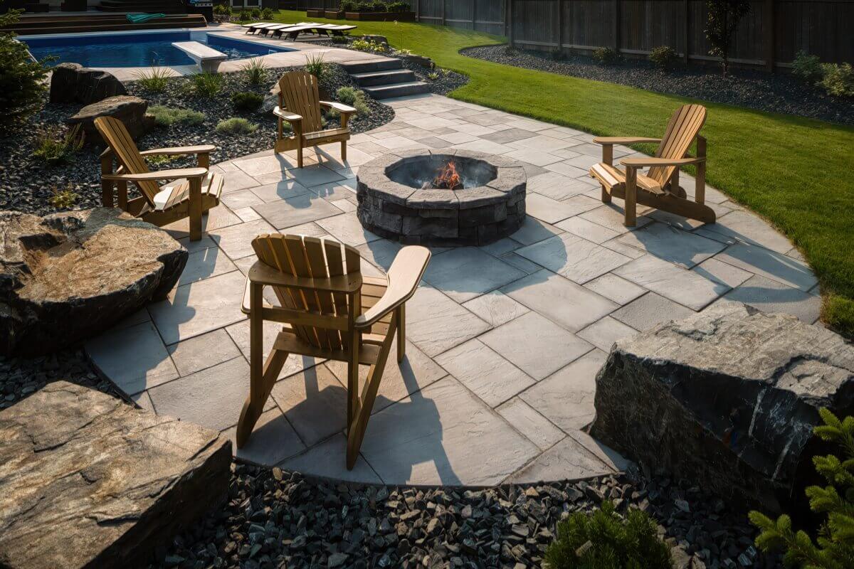 Spacious backyard setting with the Rosetta® Belvedere Fire Pit placed on a large stone patio near a swimming pool. The circular pit, made from grey and brown concrete blocks that replicate the character of weathered stone, sits at the centre of four natural-wood Adirondack chairs arranged evenly around it. Small orange flames flicker gently, casting shadows across the wide light-grey patio pavers. In the background, vibrant green grass and landscaping stones frame the peaceful atmosphere. The open space and natural textures show how the Belvedere Fire Pit creates a comfortable and generously sized area to settle in for a summer of backyard serenity.