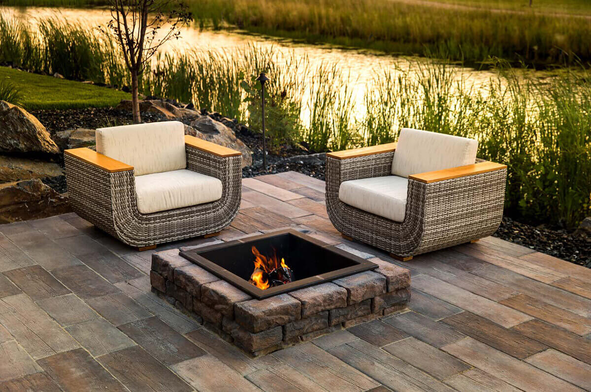 A serene outdoor patio beside a calm lake at golden hour featuring the Rosetta® Dimensional Fire Pit Kit as its centrepiece. The square fire pit is crafted from precast concrete blocks that perfectly imitate hand-chiseled natural stone, each piece showing soft variations of warm brown and charcoal grey. A matte-black steel insert frames flickering orange flames that glow against a background of reflective water and tall grass. Two rattan armchairs with cream cushions and wooden armrests sit opposite one another on a patio of rectangular pavers in earthy tones. The scene glows with evening sunlight, showing how the natural stone look of the Dimensional Fire Pit Kit transforms the patio into an inviting, warm retreat ideal for lakeside relaxation.