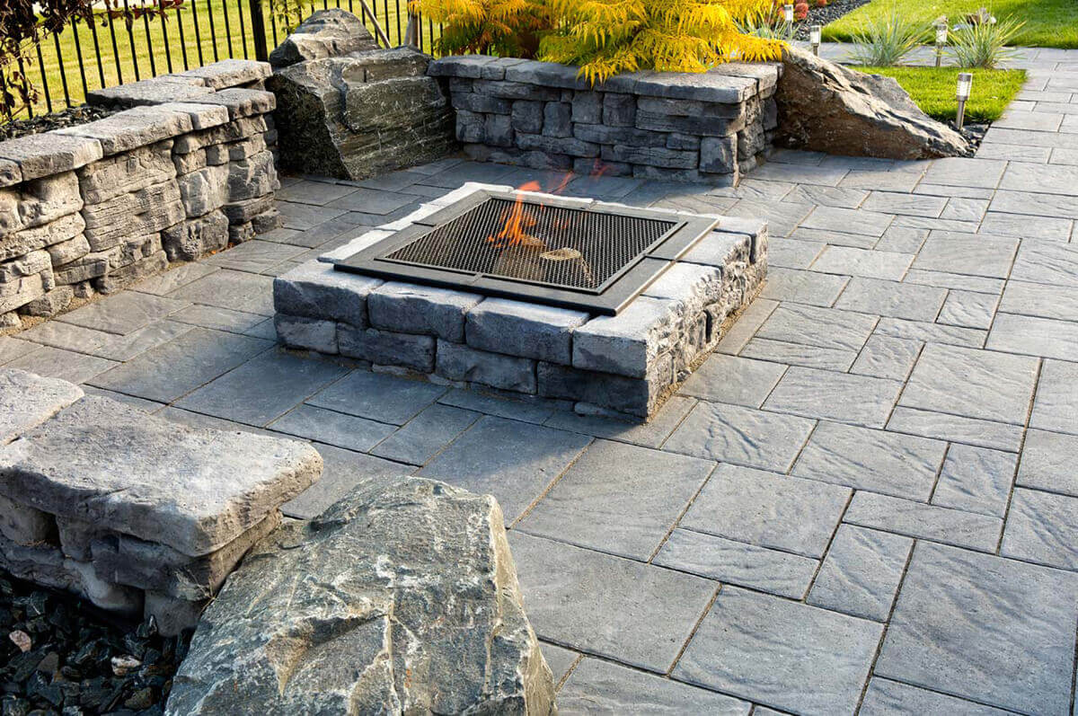 Close-up outdoor shot of a Rosetta® Dimensional Fire Pit Kit installed within a modern courtyard bordered by stone walls and landscaped greenery. The square pit is built from rugged light-grey concrete blocks, each textured to mimic split natural stone with visible grain, rough edges, and subtle shading. A black steel fire insert fitted with a diamond-mesh screen covers softly glowing embers beneath a small flame. The patio floor is made from cool-toned rectangular pavers, creating an elegant pattern beneath gentle afternoon light. The organic texture of the fire pit blocks blends harmoniously with surrounding rocks and plants, naturally showing how its stone-like finish turns the courtyard into an inviting, warm retreat.