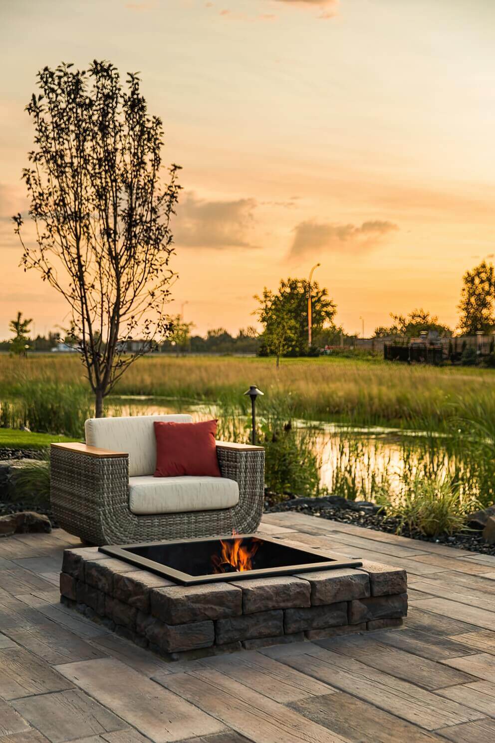 A tranquil sunset scene showcasing a single woven-rattan armchair with cream cushions and a red throw pillow facing the Rosetta® Dimensional Fire Pit Kit. The square pit, constructed from rough-faced brown-grey concrete blocks, contains a black steel insert glowing with vivid orange flames. The patio’s multi-toned pavers mirror the amber hues of the setting sun, while tall grasses and a young tree silhouette softly against the golden sky. The warm light and natural textures together highlight how the Dimensional Fire Pit Kit’s stone-inspired design transforms the lakeside patio into an inviting and peaceful retreat.