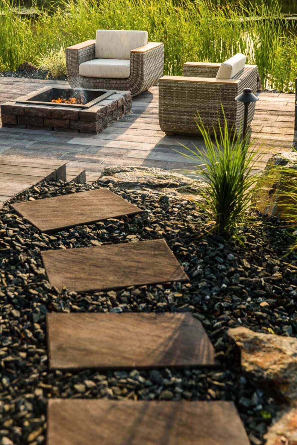 A broad late-afternoon view of a landscaped backyard patio featuring the Rosetta® Dimensional Fire Pit Kit in the foreground. The square structure is built from multi-hued brown and grey concrete blocks with a convincingly natural stone texture and irregular surface. A smooth black steel insert rests neatly inside, holding glowing firewood and low, amber-coloured flames. Two curved rattan chairs with beige cushions and wooden armrests face the fire pit, surrounded by paving stones laid in a staggered pattern. A garden path of dark stepping stones leads through gravel and tall ornamental grass toward the seating area. The craftsmanship and soft sunlight together show how the natural stone look of the Dimensional Fire Pit Kit transforms any patio into an inviting, warm retreat for gathering.