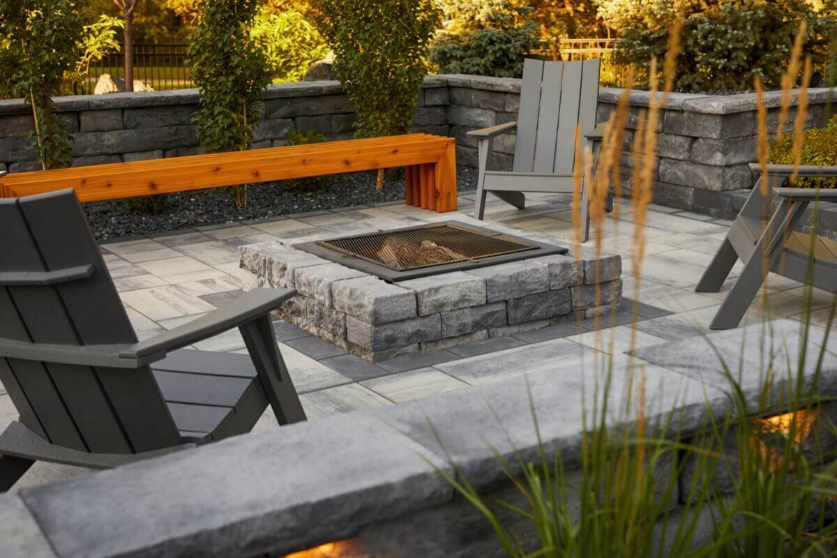 Cosy backyard courtyard setting with a Rosetta® Dimensional Fire Pit Kit at its centre, surrounded by four dark-grey Adirondack chairs and a long cedar-coloured wooden bench. The square pit is built from stacked light-grey, stone-textured blocks with slightly uneven faces that create depth and realism. A black metal mesh spark guard covers a gentle fire glowing beneath. The patio floor features large, interlocking pavers in grey and cream tones arranged in a geometric pattern, complemented by stone planters and green foliage. The warm light of early evening enhances the pit’s stone-like texture, naturally demonstrating how the Dimensional Fire Pit Kit brings an inviting warmth and retreat-like atmosphere to any backyard space.