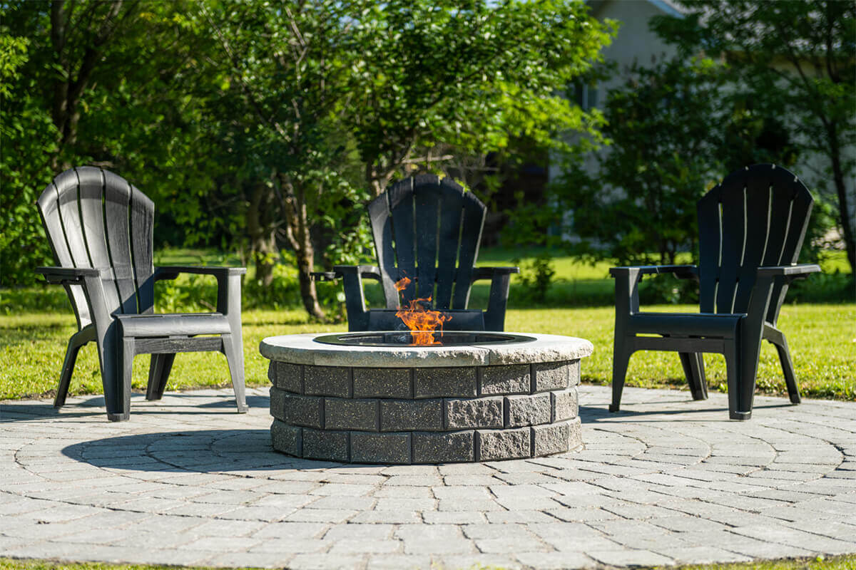 Outdoor setting featuring a Barkman StackStone® Fire Pit as the centrepiece of a circular stone patio bordered by lush green grass and tall leafy trees under bright sunlight. The round fire pit is built from two stacked layers of dark grey concrete blocks with a rough, speckled stone texture and uniform horizontal mortar lines. Each block has a slightly beveled edge that gives depth and shadow definition. A smooth, light-coloured stone coping ring sits atop the structure, contrasting with the darker base. The centre contains a black metal liner where orange and yellow flames rise gently, flickering against smouldering embers. Three matte charcoal-black Adirondack chairs surround the pit in a semi-circle, evenly spaced and facing inward. The distant greenery adds depth, creating an atmosphere of warmth and relaxation that perfectly conveys the tagline Add Warmth to Your Backyard.