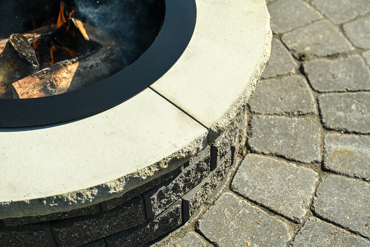 Close-up angled view of the Barkman StackStone® Fire Pit showing meticulous construction details. The image highlights the meeting point of two coping stones made of pale concrete with an unpolished, slightly rough outer edge that shows small aggregate specks. A circular black steel insert lines the inner edge, framing burning wooden logs that glow with orange firelight and produce light grey smoke. The textured concrete blocks beneath have a dual-tone surface — a blend of charcoal grey and deep brown hues — with visible rough pores and a sand-like grain pattern. The surrounding patio surface is composed of precisely cut paving stones arranged in concentric circular patterns radiating from the pit, displaying subtle wear and fine joint lines. This detailed view emphasises the weather-resistant craftsmanship and rugged charm of the StackStone® Fire Pit.