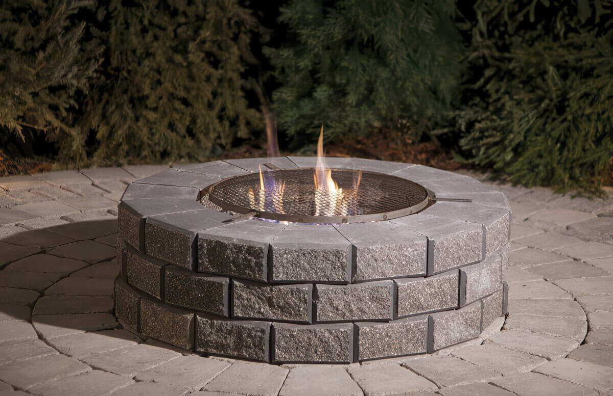 Nighttime scene of a Barkman StackStone® Fire Pit glowing against a backdrop of deep green conifer trees. The circular pit is built from stacked, square-faced grey concrete blocks with fine texture and consistent height alignment. The top ring is capped with smooth coping stones that reflect the warm glow of the fire within. A round metal mesh spark guard with a thin steel rim sits across the top, its perforated surface illuminated by small, golden flames dancing beneath it. The subtle shadows cast by the fire reveal the block contours, while the paved patio around the pit, made of large rectangular stones, gleams faintly from reflected firelight. This evening composition captures the sense of intimacy and calm associated with Add Warmth to Your Backyard.