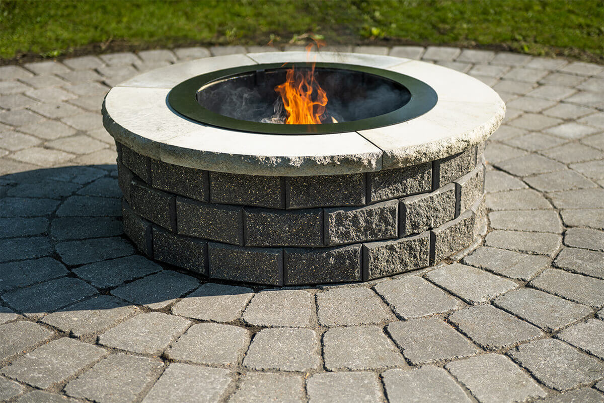 Bright daylight close-up of a Barkman StackStone® Fire Pit installed on a circular patio with concentric paving stones. The fire pit features two neat rows of dark grey, rough-textured concrete bricks arranged in perfect alignment. Each block has a distinct grain pattern, with small flecks of beige and charcoal visible across the surface. The coping ring above is made of coarse, light-beige stone, its outer edge irregular and naturally jagged, adding rustic charm. The black steel liner forms a clean inner ring, housing active orange flames producing soft, wispy grey smoke. Shadows cast by the high midday sun create crisp contrast, enhancing the fire pit’s layered texture and craftsmanship. The photo conveys the solid build and contemporary look of the StackStone® Fire Pit, embodying the spirit of outdoor warmth and durability.