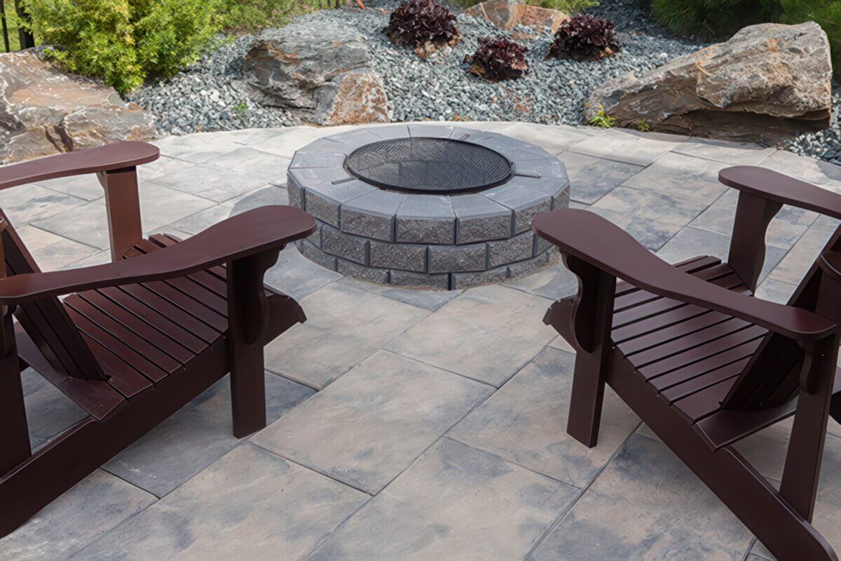 A Barkman StackStone® Fire Pit situated on a large, square-patterned stone patio framed by natural landscaping with decorative rocks and low shrubs. Two dark reddish-brown Adirondack chairs face the fire pit in the foreground, their wide armrests and curved backs catching soft daylight. The fire pit’s circular form consists of interlocking grey StackStone® bricks with lightly speckled surfaces and consistent joints, topped with a ring of coping stones in a cooler grey tone. A circular black metal mesh spark guard sits atop the pit’s centre, reflecting subtle highlights from the ambient light. Behind the setup, large textured rocks and green foliage complement the earthy palette. The composition evokes a relaxed and inviting atmosphere — a perfect visual representation of the tagline Add Warmth to Your Backyard.