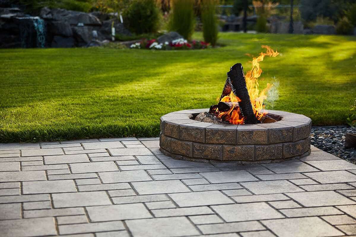 Vibrant daytime shot of a Barkman StackStone® Fire Pit positioned on a clean, geometric stone patio bordered by a vivid green lawn. The fire pit is built from light brown, speckled concrete blocks with a rough-stone texture and seamless circular alignment. Pale grey coping stones form a smooth top rim with visible grain, contrasting the earthy tones beneath. In the centre, bright orange and yellow flames rise from stacked blackened firewood, producing light smoke that dissipates into the air. The sunlight reflects off the paving stones, highlighting their rectangular pattern and fine grout lines. The background reveals manicured grass, decorative shrubs, and a rock garden with cascading water, enhancing the tranquil yet dynamic outdoor setting. The image embodies the natural beauty and warmth promoted by the tagline Add Warmth to Your Backyard.