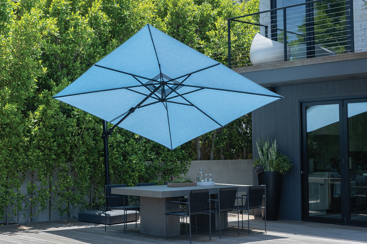 Modern patio setting featuring a bright blue AG25T Cantilever Umbrella with a wide square canopy shading a sleek grey concrete dining table. The black aluminium support pole extends from the side, anchored in a sturdy base positioned on the wooden deck. The surrounding area includes a grey exterior home with glass doors, balcony railings above, and vibrant greenery forming a backdrop of lush privacy.