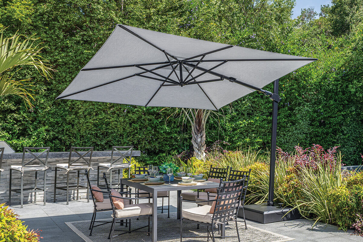 Outdoor dining area displaying a grey AG25T Cantilever Umbrella fully open above a rectangular dining table with six black-framed chairs featuring light beige cushions. The umbrella’s square canopy casts shade over a patio bordered by tall hedges and tropical plants. In the background, a raised bar area with high-back stools creates an elegant and functional backyard entertainment space.