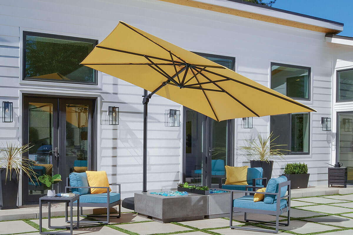 Bright courtyard scene showcasing a yellow AG25T Cantilever Umbrella shading a modern seating area with grey concrete fire table and matching furniture. The umbrella’s frame and base are dark bronze, complementing the mustard yellow canopy. Cushions in teal and gold accent the contemporary space, set against a white house façade with large windows and potted greenery.