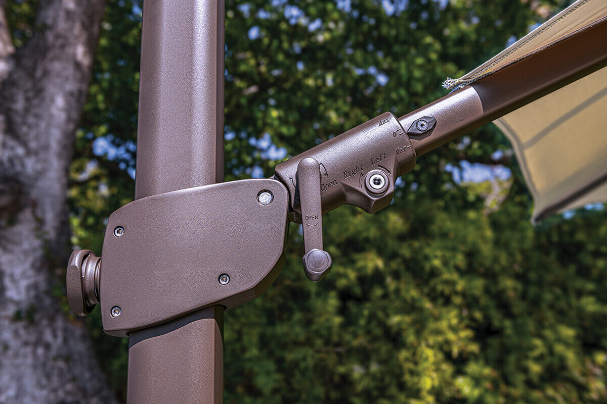 Close-up of the bronze AG25T Umbrella tilt mechanism showing the engraved direction markers for left, right, open, and max tilt angles. The precision-moulded aluminium components connect the horizontal arm to the main mast with visible bolts and a smooth matte metallic finish. Sunlight reflects off the bronze surface, highlighting the umbrella’s mechanical quality and durable construction.