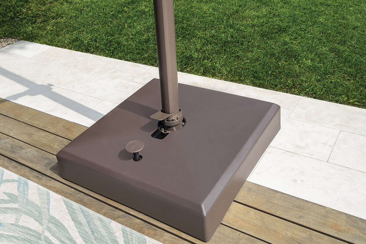 Detailed top-down view of the bronze AG25T Umbrella base on a wooden deck. The square resin base securely anchors the umbrella mast with a rotating foot pedal and locking lever. The setting includes neatly edged grass and pale stone tiles nearby, accentuating the umbrella’s stability and refined design suited for upscale patios.