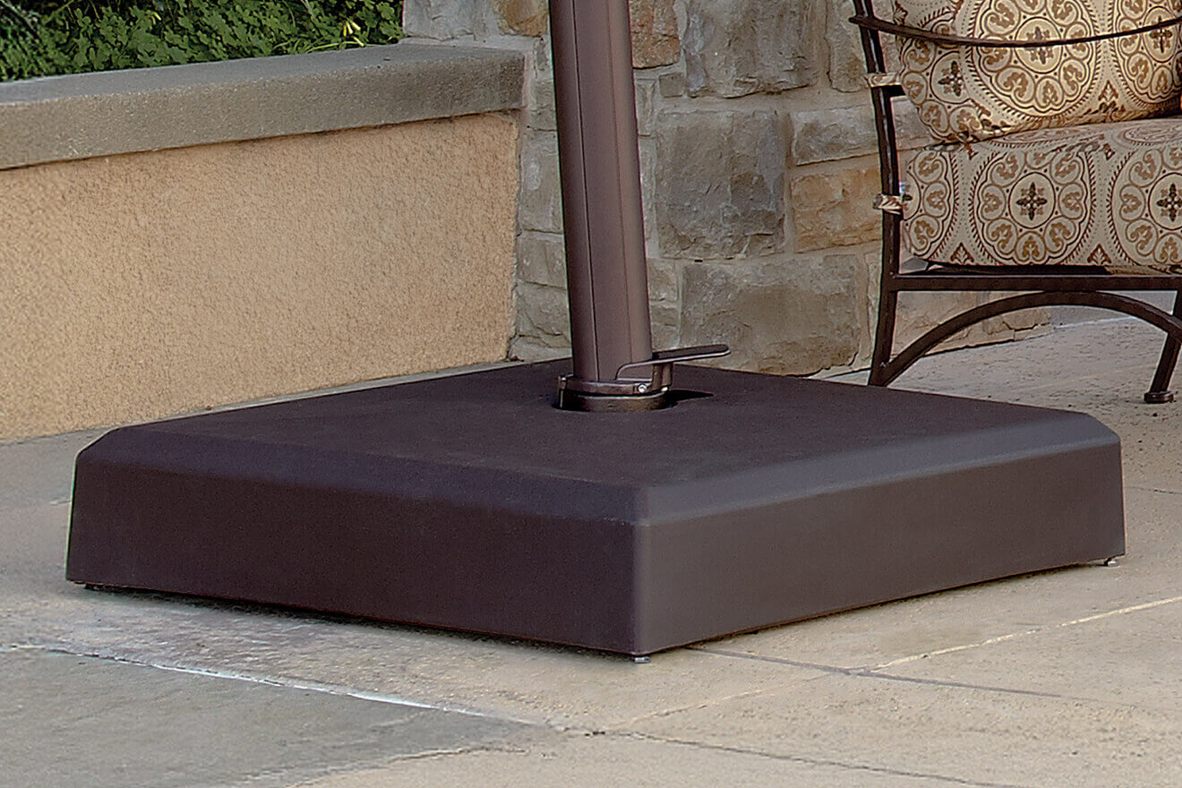 Outdoor view of the AKZ Sand-Filled Resin Base in bronze finish, securely holding a cantilever umbrella pole on a textured stone patio beside a cushioned outdoor chair with a decorative beige and brown pattern. The low, square base has a smooth surface with a central circular opening for the umbrella post, its edges softly bevelled for a streamlined appearance. The bronze powder-coated steel frame blends into the patio setting, offering a stable, weather-resistant foundation for the umbrella.